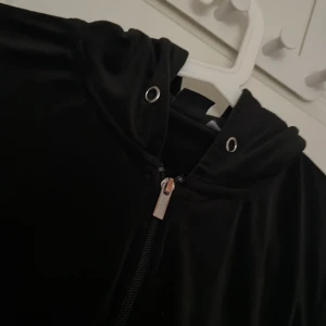Svart hoodiejacka med dragkedja och byxor  - Säljer en svart kofta med dragkedja framtill och huva. Koftan har en slät yta och är enkel i modellen och ett par matchande mjukisbyxor , perfekt för en avslappnad stil. Passar bra till vardags och är lätt att matcha med andra plagg. Storlek M i koftan och S i byxorna  ( bandet till både luvan och byxorna saknas ) 