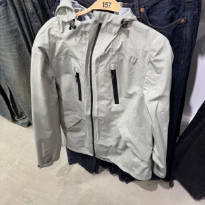 Skaljacka - Säljer en ljusgrå vindjacka i storlek XL. Jackan har huva, dragkedja framtill, två bröstfickor med dragkedja och två sidofickor. Justerbar nederkant och ärmslut med reflexdetaljer. Perfekt för blåsiga dagar.