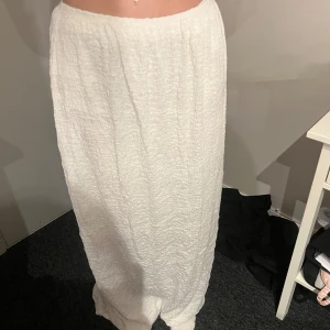 Vit långkjol i krinklat tyg - Säljer en vit långkjol med krinklad struktur och resår i midjan. Kjolen har en rak passform och är tillverkad i ett lätt material som passar perfekt till varmare dagar.