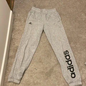 Grå mjukisbyxor från Adidas - Grå mjukisbyxor från Adidas med resår i midjan och muddar vid bensluten. Byxorna har Adidas-logga på ena benet och är tillverkade i mjukt bomullsmaterial. Perfekta för träning eller chill.