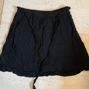 Svart kortkjol från Vero Moda, S - Svart kortkjol från Vero Moda i mjukt tunnt bomullstyg. Kjolen har resår i midjan och knytband för extra passform. Enkel och stilren design som är lätt att matcha med allt. Perfekt för sommaren! Använt kanske 2 gånger