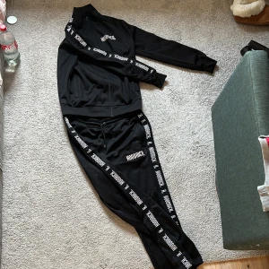 Svart tracksuit från Hoodrich - Säljer en svart tracksuit från Hoodrich med tydliga loggor längs benen, armarna och på höften. Byxorna har resår i midjan och är tillverkade i ett mjukt syntetmaterial. Perfekta för en avslappnad streetwear-look. Tröjan har en dragkedja och detaljer längs ärmarna. Tröjan är i storlek Medium och byxorna Large. Skriv om ni vill köpa separat.