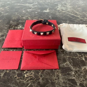 Valentino armband - Säljer ett svart armband från Valentino Garavani med ikoniska pyramidformade silverfärgade nitar. Armbandet är gjort i skinn och har ett justerbart spänne. Kommer med original ask, dustbag och äkthetskort. Perfekt accessoar för en edgy look. Det är hundra procent äkta!
