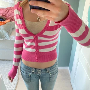 Randig kofta - Säljer en söt kofta med rosa och vita ränder. Modellen är croppad med långa ärmar och knappar framtill. Perfekt för dig som gillar färg och vill ha en stickad tröja med lite edge. Liten defekt på bröstet, se bild 4💕