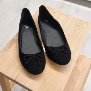 Svarta ballerinaskor med rosett - Säljer ett par klassiska svarta ballerinaskor med rund tå och söt rosettdetalj framtill. Perfekta att matcha till både jeans och klänning. Enkla att ta på och av, och passar till många olika stilar.
