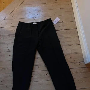 Svarta kostymbyxor från Nelly - Stilrena svarta kostymbyxor från Nelly med klassisk rak passform och hällor för bälte. Byxorna har diskret dragkedja och knapp framtill samt sidfickor. Perfekta för en clean och dressad look.