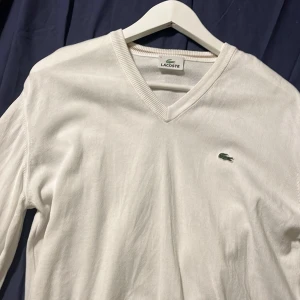 Vit v-ringad tröja från Lacoste - Vit v-ringad tröja från Lacoste med klassisk krokodillogga broderad på bröstet. Tröjan är långärmad och har ribbade muddar vid hals, ärmslut och nederkant. Tillverkad i mjukt bomullsmaterial, perfekt för en clean och stilren look. Storlek M