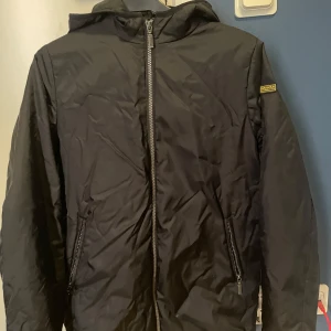 Svart vindjacka barbour - Svart vindjacka med huva från barbour. Jackan har dragkedja framtill och två snedställda fickor med dragkedja. Enkel och stilren design, perfekt för blåsiga dagar.
