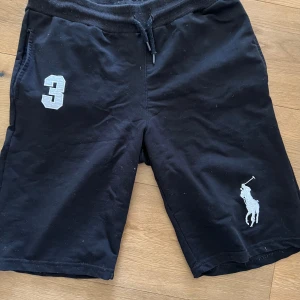 Svarta shorts från Polo Ralph Lauren - Svarta shorts från Polo Ralph Lauren med snörning i midjan. Stort vitt Polo-logga broderat på ena benet och en vit siffra 3 på andra benet. Sköna och sportiga, perfekta för sommaren.