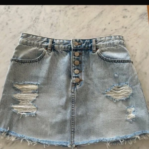 Ljusblå jeanskjol med slitningar och knappar - Säljer en ljusblå jeanskjol med slitna detaljer och rå kant nertill. Kjolen har knappar framtill, klassiska fickor både fram och bak samt bälteshällor. Perfekt för en avslappnad och trendig look.