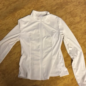 Vit sportig vindjacka med dragkedja - Snygg vit vindjacka med hel dragkedja framtill och långärmad design. Jackan har en sportig look med figurnära passform och hög krage. Materialet är lätt och känns syntetiskt, perfekt för aktiva dagar. Enkel och stilren utan mönster eller extra detaljer.
