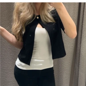 Svart cardigan blus med knapp - Kort svart cardigan blus med en snygg guldig knapp upptill. Tröjan har korta ärmar och en öppen front, perfekt att slänga över ett linne eller t-shirt för en clean och stilren look.🥰