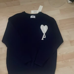 Mörkblå stickad tröja från Ami Paris  - Mörkblå stickad tröja från Ami Alexandre Mattiussi (Ami Paris) med vit hjärtlogga och bokstaven A på bröstet. Klassisk rund halsringning och långa ärmar. Perfekt för dig som gillar stilrena och trendiga plagg med en twist.