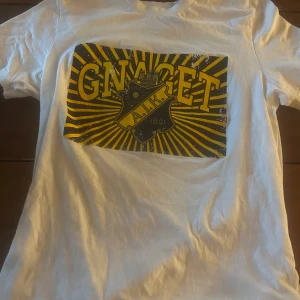 Vit AIK t-shirt med GNAGET-tryck - Vit t-shirt från AIK med stort gult och svart GNAGET-tryck på bröstet och klubbens emblem. Klassisk passform och rund halsringning. Perfekt för dig som vill visa ditt AIK-stöd på läktaren eller i vardagen.