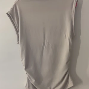 Beige ärmlös topp med rynkade sidor - Säljer en stilren beige topp med ärmlös design och snygga rynkade detaljer längs sidorna. Toppen har en mjuk och slät yta, perfekt för dig som gillar minimalistisk stil. Passar till många olika outfits och är superenkel att matcha.