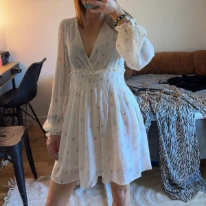 Vit blommig kortklänning med v-ringning - Supersöt vit kortklänning med små blommor, v-ringad och smockad midja. Långa, luftiga ärmar och lätt, flowy material. Perfekt för sommarens alla tillfällen! Ny med prislapp 