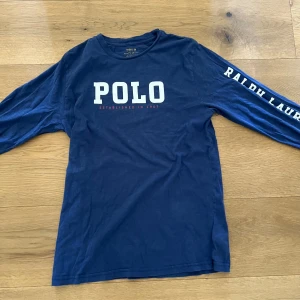 Blå långärmad tröja från Polo Ralph Lauren - Säljer en blå långärmad tröja från Polo Ralph Lauren med stor vit POLO-text på bröstet och logga på ärmen. Tröjan har rund hals och är tillverkad i mjuk bomull. Perfekt för en avslappnad stil.