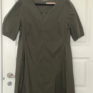 Olivgrön klänning med puffärm - Snygg olivgrön klänning med v-ringning och puffiga korta ärmar. Klänningen har en rak passform och är tillverkad i ett mjukt bomullstyg. Perfekt för dig som gillar stilrena och trendiga plagg.