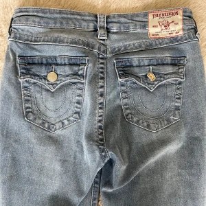 True Religion Becca Mid Rise Bootcut Jeans - Snygga ljusblå jeans från True Religion, modell Becca med mid rise och bootcut. Klassiska bakfickor med dekorativa sömmar och silverfärgade knappar. (En knapp på ena fickan har lossnat och vikt sig i tvätten) ANVÄNDA 1 GÅNG/ NYPRIS 1299