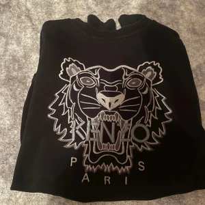 Kenzo tröja - Säljer en svart sweatshirt från Kenzo med ett stort broderat tigerhuvud och texten 'Kenzo Paris' på framsidan. Tröjan har långa ärmar och ribbade muddar vid ärmslut och nederkant. Perfekt för en stilren look.