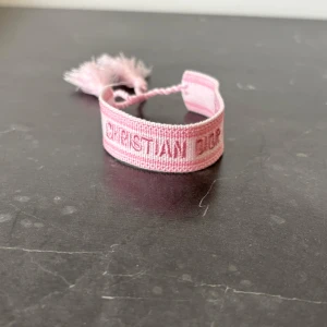 Rosa armband från Christian Dior - Säljer ett ljusrosa armband från Christian Dior med broderad logga och två tofsar. Armbandet är justerbart med flätad knytning och har en mjuk textilstruktur. Perfekt accessoar för att lyfta din outfit.