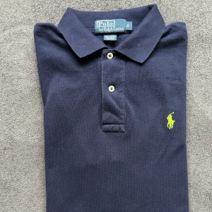 Ralph Lauren piké mörkblå - Mörkblå pikétröja i S från Ralph Lauren med gul broderad logga på. Skick 7/10 om inte mer, smått urtvättad om något men inget som syns. Hör av er vid frågor!