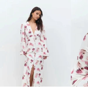 Vit blommig långklänning med v-ringning - Säljer en vit långklänning med rosa blommigt mönster, djup v-ringning och långa ärmar. Klänningen har knappar framtill och en slits som ger en snygg detalj. Perfekt för sommarens tillfällen! Storlek 36.