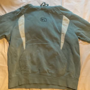 Nike Air hoodie i blågrönt - Nike Air hoodie i blågrönt med vita detaljer längs sidorna och klassisk logga på bröstet. Tröjan har huva, stor magficka och långa ärmar. Perfekt för en chill och sportig stil. Mjuk och skön känsla tack vare bomullsmaterialet. Byxor följer med