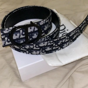Mönstrat bälte  - Säljer ett snyggt bälte från  med blått och vitt mönster samt en svart metallspänne med CD-logga. Bältet har en exklusiv känsla och passar perfekt till jeans eller kostymbyxor.