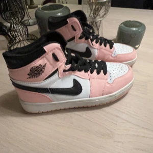Nike Air Jordans i rosa och svart - Snygga Nike Air Jordans i en fräsch kombination av rosa, svart och vitt. Skorna har en hög modell med klassisk snörning och den ikoniska Nike-loggan på sidan. Perfekta för att ge din outfit en sportig touch.