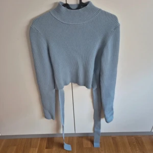 Ljusblå ribbad tröja med knytband - Snygg ljusblå ribbad tröja med hög krage och långa ärmar. Tröjan har en unik design med knytband som ger en cool look. Perfekt för att uppdatera garderoben med något stilrent och trendigt.