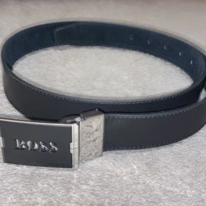 Svart bälte från Hugo Boss - Snyggt svart bälte från Hugo Boss med en stilren silverfärgad spänne där 'BOSS' är ingraverat. Perfekt för att ge en elegant touch till din outfit. Bältet är gjort av skinn och har flera justeringshål för bästa passform.