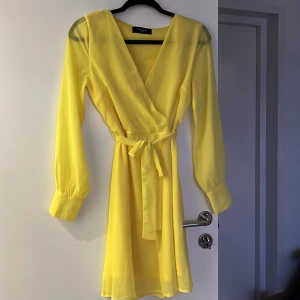Gul omlottklänning från Sisters Point - Snygg gul omlottklänning från Sisters Point med knytband i midjan. Klänningen har långa ärmar och en v-ringad design som ger en elegant look. Perfekt för att lysa upp garderoben med en färgglad touch! 