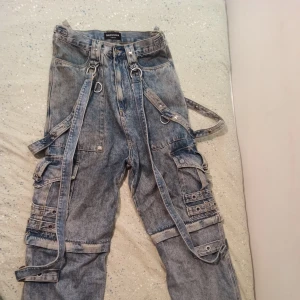 Raver Jeans - Unika blå jeansbyxor med flera spännande detaljer som dragkedjor och remmar. Byxorna har en loose passform och är dekorerade med metallringar och spännen. Perfekta för en edgy look.