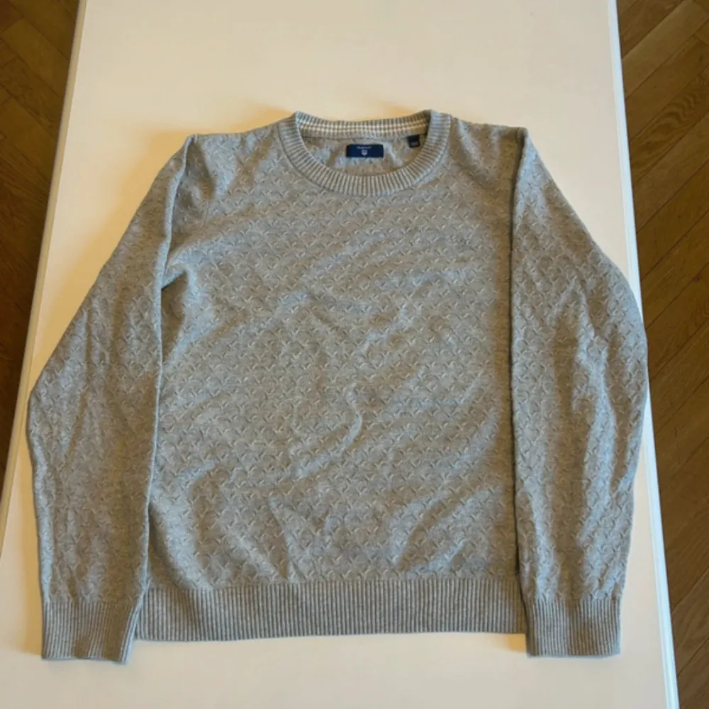 Säljer en stilren grå tröja från GANT med ett diskret mönster. Tröjan har långa ärmar och ribbade muddar vid ärmslut och nederkant. Perfekt för en avslappnad och snygg look.. Neuletakit & Villapaidat.