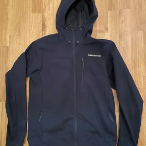 Mörkblå jacka från Peak Performance - Snygg mörkblå jacka från Peak Performance med huva och dragkedja. Jackan har flera praktiska fickor med dragkedjor och är perfekt för utomhusaktiviteter. Den är tillverkad i ett slitstarkt material och har en stilren design.