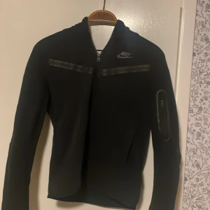 Svart jacka från Nike - Säljer en svart jacka från Nike med dragkedja och huva. Jackan har långa ärmar och en stilren design med Nike-logga på bröstet. Perfekt för en sportig look. Köpte för 1500 på Jd Sports, välanvänd men fortfarande i bra skick