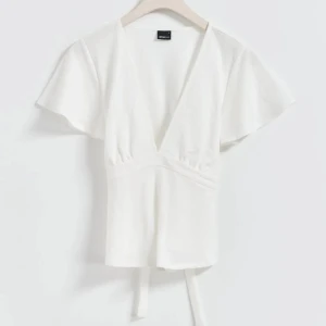 Vit omlottopp med knytband - Säljer en stilren vit omlottopp med korta ärmar och knytband i midjan. Toppen har en v-ringad design som ger en elegant touch. Perfekt för en avslappnad men chic look.