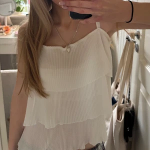 Vit plisserad topp - Säljer en söt vit plisserad topp med volanger. Toppen har smala axelband och en luftig passform, perfekt för varma dagar. Den är stilren och lätt att matcha med olika outfits. Perfekt för en somrig look! 🌸