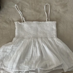 Vit topp från Gina Tricot - Säljer en söt vit topp från Gina Tricot med smockdetaljer upptill och tunna axelband. Den har en luftig och fladdrig nederdel som ger en romantisk känsla. Perfekt för varma sommardagar!