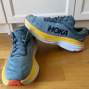 Hoka Bondi 8 löparskor - Säljer ett par Hoka Bondi 8 löparskor i en snygg kombination av blått, gult och rött. Skorna har en tjock sula för extra dämpning och komfort. Perfekta för långa löprundor med bra stöd och stabilitet.