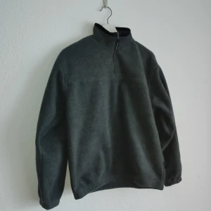 Grå fleece tröja från Clique - Mysig grå fleece tröja från Clique med halv dragkedja och hög krage. Perfekt för kyliga dagar med sin mjuka och varma känsla. Tröjan har långa ärmar och en enkel design som passar till det mesta.