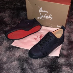 Svarta sneakers från Christian Louboutin - Snygga svarta sneakers från Christian Louboutin med den ikoniska röda sulan. Skorna har en stilren design med snörning och är tillverkade i mocka. Perfekta för den som vill ha en lyxig touch i vardagen.