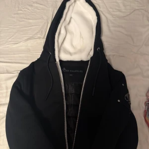 Svart jacka med vit foder från Moose Knuckles - Säljer min moose knuckles jacka/ hoodie perfekt inför våren säljer pga jag växte ur den direkt. Knappt använd som man ser på bilderna kvitto finns och den är i nyskick 