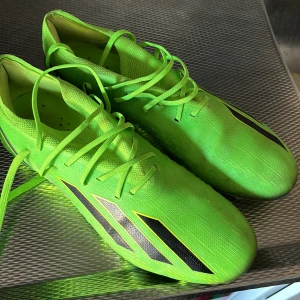 Gröna fotbollsskor från Adidas - Snygga gröna fotbollsskor från Adidas med svarta detaljer och snörning. Skorna har en modern design med dobbar för bra grepp på planen. Perfekta för fotbollsspelare som vill sticka ut på planen.