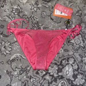 Rosa glittrig bikini från Roxy - Snygg rosa bikini från Roxy med glittriga detaljer. Överdelen har trekantsform och knytband runt nacken och ryggen. Underdelen har knytband på sidorna för justerbar passform. Perfekt för sommarens stranddagar!