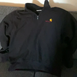 Svart halfzip från Carhartt - Säljer en svart tröja från Carhartt med halv dragkedja och broderad logga på bröstet. Tröjan har långa ärmar och en hög krage, perfekt för kyligare dagar. Passar både till vardags och mer avslappnade tillfällen. Passar medium/large ,,, nypris 1299:-