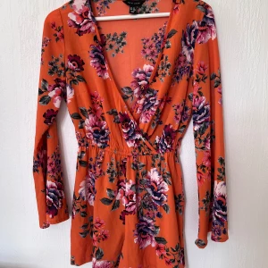 Orange blommig jumpsuit  - Snygg orange jumpsuit med blommigt mönster i rosa och lila nyanser. Klänningen har en omlott-design med v-ringning och långa ärmar. Perfekt för en färgglad look!