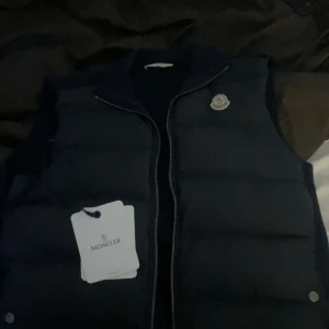 Mörkblå dunväst från Moncler - Säljer en snygg mörkblå dunväst från Moncler. Västen har en dragkedja framtill och är perfekt för kyligare dagar. Den har en ribbad krage och Monclers logga på bröstet. Passar perfekt för en stilren look. Det är både tun och dunväst