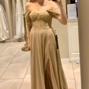 Champagnefärgad långklänning med spetsdetaljer - Endast använd 1 gång. Elegant champagnefärgad långklänning med spetsdetaljer på överdelen och en vacker slits. Klänningen har offshoulder-ärmar och en omlott-design som ger en romantisk känsla. Perfekt för speciella tillfällen. Inköpt för 5000kr på Amará Stockholm Brud, fest & balklännings butik förra året. Uppsydd, jag är 162cm och hade 8 centimeters klackar då nådde den precis till golvet. 
